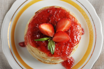 On a plate of pancakes, poured with strawberry jam and decorated with fresh strawberries. The background is draped with a napkin. View from above. Close-up. Macro photography.