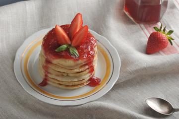 On a plate of pancakes with strawberry jam, decorated with fresh strawberries. The background is draped with a napkin. In the frame, a strawberry, a spoon, a jar of jam. Close-up. Macro photography.