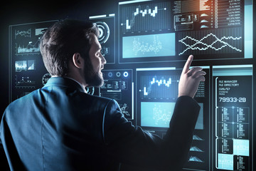 Amazing technologies. Clever emotional young programmer pointing to the transparent screen of a futuristic device and feeling impressed by the power of modern technologies