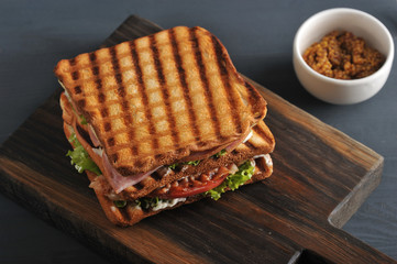 Sandwich of several slices of white bread on a wooden board. The filling of the sandwich consists of bacon, cheese, ham, sauce, tomato and lettuce. Next cup with mustard. Dark background. Close-up.