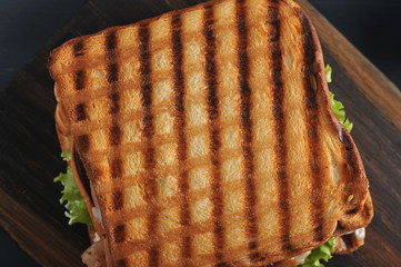 Sandwich of several slices of white bread toasted on the grill. The filling of the sandwich consists of bacon, cheese, ham, sauce, tomato and lettuce. Dark background. Close-up. Macro photography.