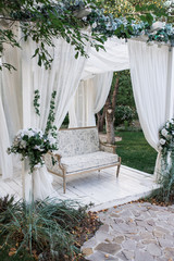 Fototapeta na wymiar In the garden there is a podium on which a beautiful white sofa in the style of Provence or rustic. Above the sofa is an arch with blossoms and flowing white fabric. Decoration for a wedding, scenery