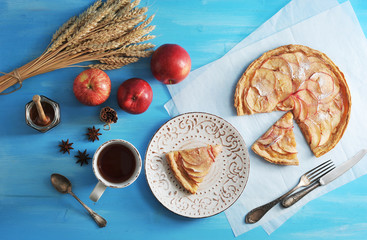 Traditional French pie is apple tartar. A piece of cake is cut off. Next a cup of tea and a piece of cake on a plate. Several apples, a jar of honey, wheat ears, anise and cinnamon. View from above. 