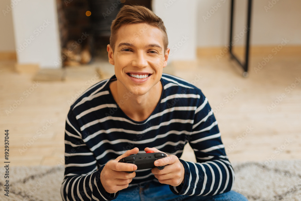 Wall mural Beaming. Good-looking content fair-haired young man smiling and holding a remote control for games while sitting on the floor