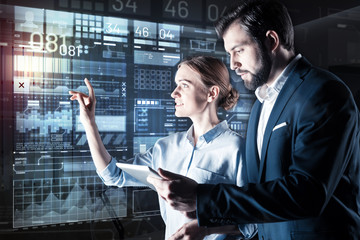 Team work. Optimistic smart woman pointing to the screen of a futuristic computer while her attentive serious colleague checking the information in his device