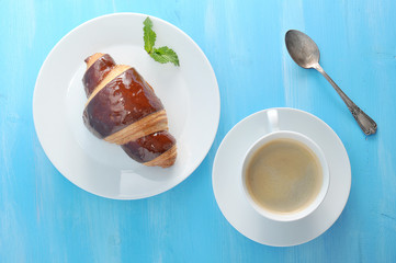 A lush and airy chocolate croissant and a cup of black coffee are the perfect combination for breakfast or a snack. On a plate with a croissant a mint leaf. Blue wood background. View from above.