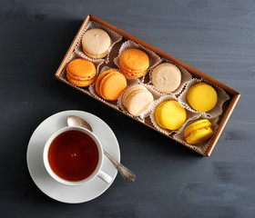 Box with multi-colored macaroons - classic French pastries based on ground almonds. Next cup with tea on a saucer. Dark wooden background. View from above. Close-up.