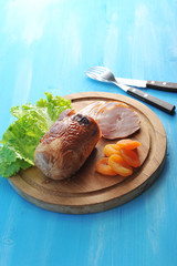 Roll of poultry meat on a wooden tray. The dish is decorated with green salad and dried apricots. There are cutlery nearby. Blue wood background. Vertical orientation of the frame.