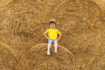 Naklejka na ściany i meble Little nice boy dressed in a yellow shirt and denim shorts sits on a haystack in a summer day. Harvesting.