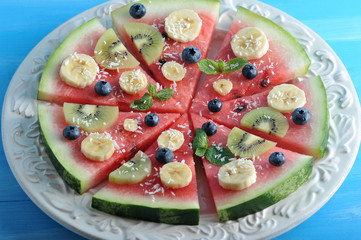 Slices of watermelon on a white plate form a berry-fruit cake. Slices of watermelon are decorated with pieces of banana, kiwi, blueberries and mint leaves. Blue wood background. 