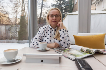 Beautiful adult woman with blond hair and glasses is waiting for a business meeting in a cafe with a white interior.