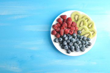 On a white plate, blueberries, raspberries and kiwi slices. Blue wood background. Free space under the text.  View from above.
