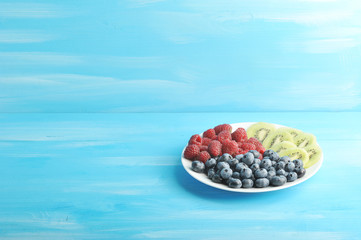 On a white plate, blueberries, raspberries and kiwi slices. Blue wood background. Free space under the text.