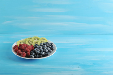 On a white plate, blueberries, raspberries and kiwi slices. Blue wood background. Free space under the text.