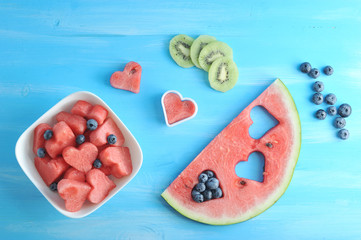 Pulp of watermelon in the shape of hearts and blueberries in a white plate. Next slices of kiwi, blueberry, a slice of watermelon, a heart shaped form. Blue wood background. View from above. Close-up.