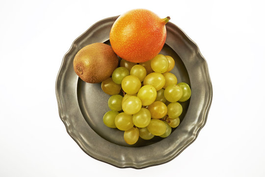 Kiść zielonych winogron, granadilla i kiwi na metalowym talerzu, centralnie z g&oacute;ry na białym tle