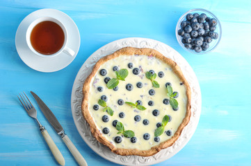 Chocolate tart with blueberries. Baking is decorated with mint leaves. Nearby a cup of tea, cutlery and a bowl of blueberries. Blue wood background. View from above. Close-up.