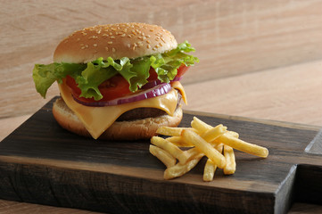 Hamburger and French fries on a wooden board. Hamburger consists of juicy meat cutlets, red onions, tomato slices, green salad, cheese. Mayonnaise and ketchup complement the taste. Close-up.