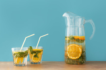 two glass transparent glasses filled with water with mint lemon, orange and straws near a glass decanter with water mint lemon and orange on a wooden table and blue background