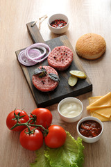 On the wooden board cutlets from minced meat, slices of pickled cucumber, onion rings. Nearby tomatoes, cheese, lettuce, bun and cups with sauces. Vertical orientation of the frame.
