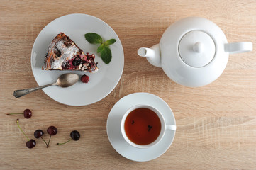Classic chocolate brownie on a plate. Brownie is decorated with a leaf of mint. Next a cup of tea and a dish of pie. Fresh cherries are on the table. Serving for one person. View from above. Close-up.