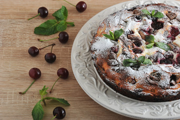 Chocolate brown curd with cherry. The cake is decorated with sugar powder and fresh mint. Fresh cherries are laid out nearby. Light wooden background. Close-up. Macro photography.