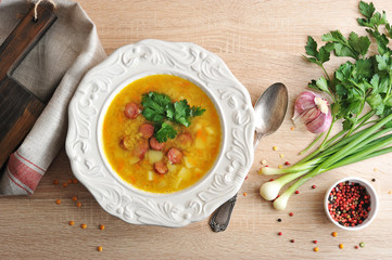 Pea soup with smoked meat in a plate. Fabric napkin. Next to the plate, a spoon and spices: garlic, parsley, green onions and peppers.  Light wooden background. View from above.