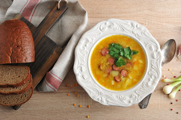 Pea soup with smoked meat in a white plate. Near the bread in a sliced on a wooden board and cloth napkin. View from above.