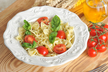 Pasta with pesto sauce and cherry tomatoes. The pasta is decorated with fresh basil leaves. Next to the dish is a jug of olive oil and a twig of tomato. Light wooden background. Macro photography.