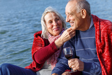 Happy senior couple enjoying time together drinking wine by the lake wrap around in a red blanket.