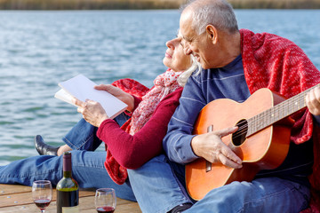 Happy senior couple enjoying time together playing guitar and drinking wine by the lake wrap around in a red blanket.