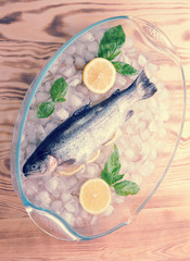 Fresh trout on ice slices. In the trouser trout a few slices of lemon. Next to fresh basil and two halves of lemon. Light wooden background. View from above. Close-up. Vertical shot.