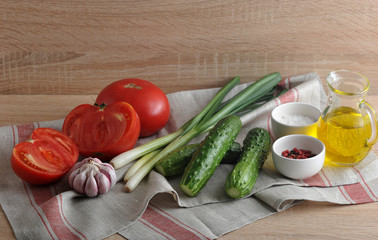 Red tomatoes, green onions, cucumbers and a garlic head on a tissue napkin. In the cups salt and pepper. Olive oil in a glass jug. Set for vegetable salad. Wooden beige background.