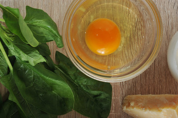 A glass bowl with a raw egg without a shell. Next to the greens of spinach and Parmesan cheese. View from above. Close-up.