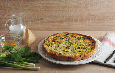 Open pie of quiche with spinach and spring onions on a white plate. Next to the jug of glass with cream, Parmesan cheese, green onions, spinach greens, glass bowl with raw eggs without shell.