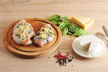 Chicken breast with ricotta cheese, curry, spinach and  on a wooden tray. Two pieces in raw  form for cooking in the oven. Nearby cheese ricotta on a white plate, greens spinach and Parmesan cheese. 