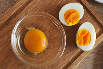 On a wooden tray are two halves of an hard-boiled eggs and a glass bowl with a raw egg without a...