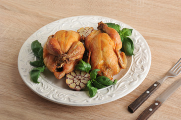 Two chicken cooked on the grill. Chickens on a white dish, next to the basil greens and halves of garlic. Cutlery: fork and knife. Fresh wooden background.