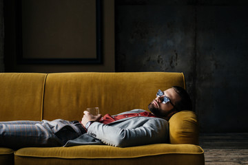 drunk man in sunglasses with glass of cognac lying on yellow sofa in loft interior