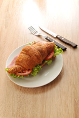 Croissant with a stuffing on a white plate. As stuffing ham, green salad, tomato and cheese. There are cutlery nearby. Light wooden background. Close-up. Vertical orientation of the frame.