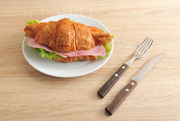 Delicious French breakfast-croissant with filling. As stuffing ham, green salad, tomato and cheese. There are cutlery nearby. Light wooden background. Close-up.