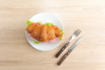 Delicious French breakfast-croissant with filling. As stuffing ham, green salad, tomato and cheese. There are cutlery nearby. Light wooden background. View from above. Close-up.