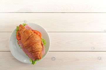 Croissant with ham filling, green salad, tomato and cheese. Light wooden background. Free space for text.