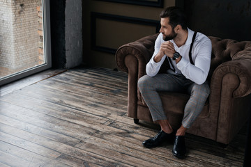 handsome stylish man sitting in armchair and looking at window, loft interior - obrazy, fototapety, plakaty