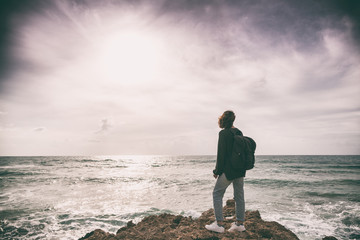 A girl with a backpack is standing with her back on the stormy seashore, an image with retro toning