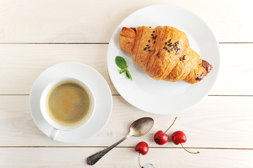 A cup of black coffee and a plate with a croissant. View from above. The croissant is decorated with chocolate crumbs. On a plate of mint leaves. Complement the composition of a teaspoon and cherry.