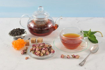 A teapot with black tea and a cup of tea. Glass ware. Next to the plate are dried rosebuds, used for brewing tea. In the frame, welding, mint, moth and crystalline sugar. Light background. Close-up.