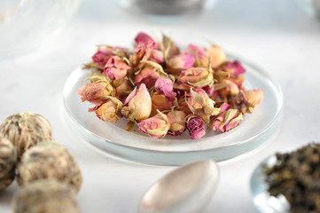 Dried buds of roses on a plate. In the background, green tea in balls. In the frame, a teaspoon. Light background. Close-up. Macro photography.