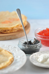 Pancakes thin on a round wooden tray and on a plate. There are two cups with red and black caviar. There is sour cream on the saucer. Light background. Vertical orientation of the frame.