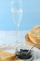 Thin big pancakes on a wooden tray. Next to the plate are two pancakes and a cup of black caviar. In the frame - a teaspoon and a glass of  white wine. Vertical orientation of the frame. Close-up.